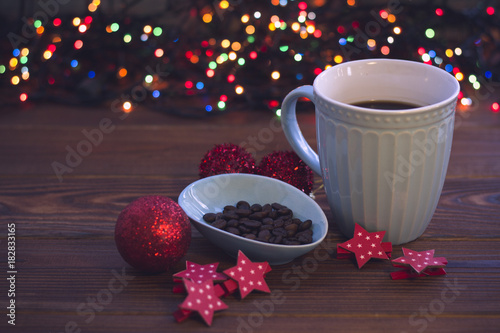 A romantic evening still life with a light blue porcelian cup of hot coffee, a bowl of coffee beans, red stars and balls against a rustic wooden background with Christmas lights. Dark colorful bokeh. photo