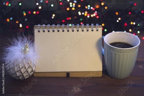 A romantic evening still life with a light blue porcelian cup of hot coffee, a blank notebook and a white and gold shabby Christmas-tree ball against a rustic wooden background with Christmas lights photo