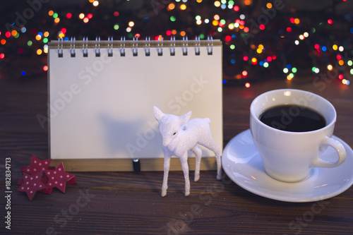 A festive still life with a balnk notebook, a cup of hot black coffee, red star clips and a toy baby deer. A white porcelian cup with a saucer. A rustic wooden background with Christmas lights. Dark photo