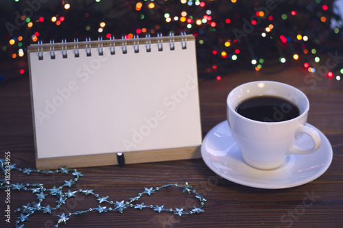 A festive still life with a balnk notebook, a cup of hot black coffee and a chain of star shaped blue beads. A white porcelian cup with a saucer. A rustic wooden background with Christmas lights. Dark photo