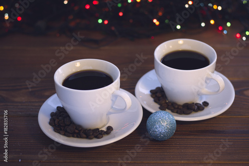 A festive still life with two white porcelian cups of hot black coffee. Coffee beans scattered on the saucers. A glitter blue Christmas-tree ball in a close up. A rustic wooden background with photo