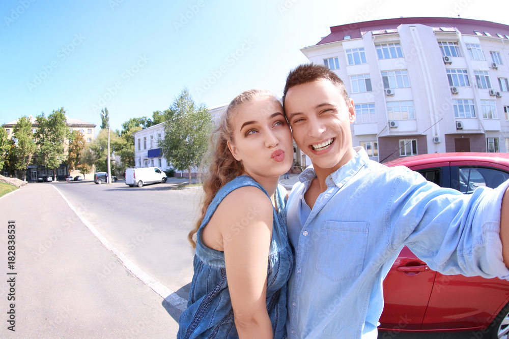Happy young couple taking selfie near new car outdoors