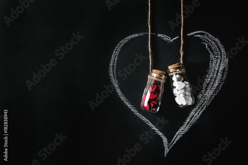 Two bottles full of white and red decorative hearts inside drawn by chalk heart shape over black background. St Valentine concept. Copy space