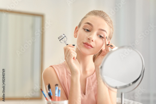 Beautiful young woman using eyelash curler at home