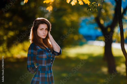 Young romantic woman in long checkered blue dress over background autumn portrait. Pretty girl posing in park with cup of coffe. Fashionable long hair model in elegant autumn clothes. Copy space