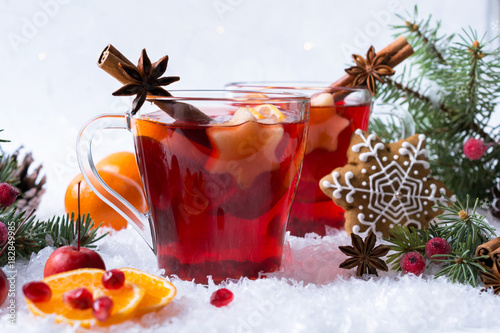 Hot mulled spiced red wine in glass mug with with festive decoration lights and gingerbread . On white snow background