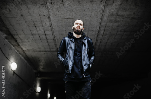Modern young bearded man in black style clothes standing in around urban background.