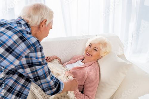 senior man covering happy wife with blanket on cozy couch