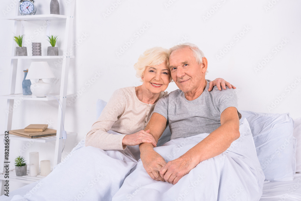 portrait of smiling senior couple resting in bed and looking at camera at home