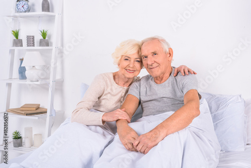 portrait of smiling senior couple resting in bed and looking at camera at home