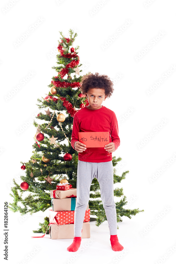 Portrait of cute mixed race little boy standing near Christmas tree holding letter to Santa