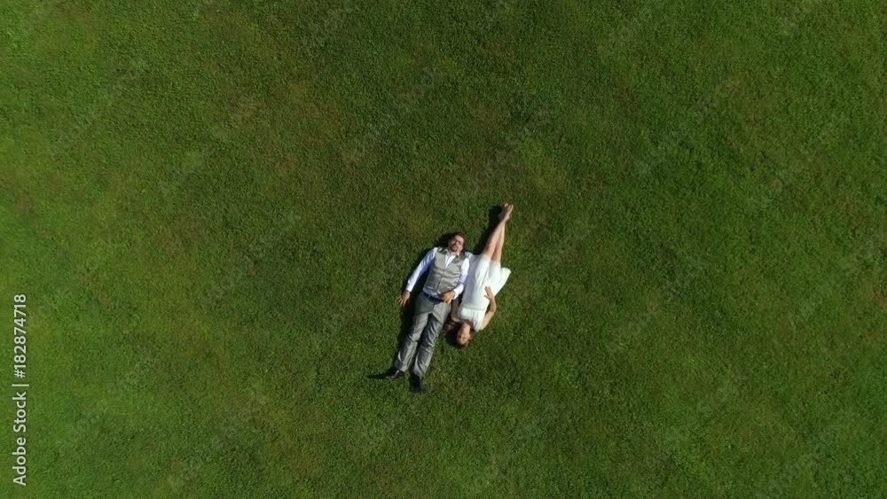AERIAL TOP DOWN: Happy young couple laying in grass on summer day. Man and wife newlyweds relaxing in sunny park after wedding. Young man and pregnant woman lying and holding hands, expecting a baby AERIAL TOP DOWN: Happy young couple laying in grass on summer day. Man and wife newlyweds relaxing in sunny park after wedding. Young man and pregnant woman lying and holding hands, expecting a baby
