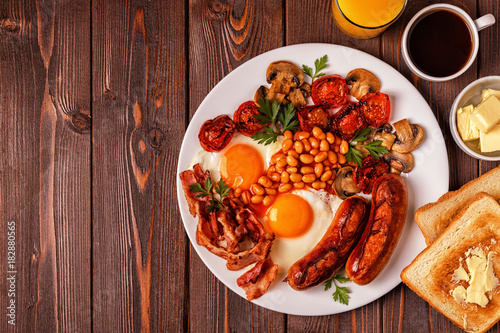 Traditional full English breakfast with fried eggs, sausages, beans, mushrooms, grilled tomatoes and bacon on wooden background.
