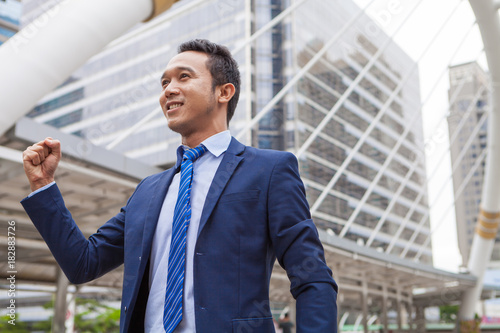 Businessman smiling and raising his fist in the air, with office building background - business success, achievement, and win concepts