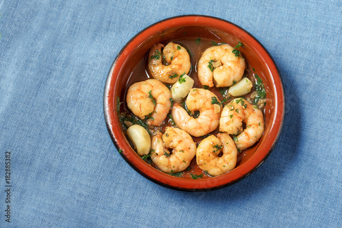 shrimps or prawns and garlic in olive oil with parsley in a brown tapas bowl, spanish appetizer on a blue tablecloth background, flat top view from above, copy space