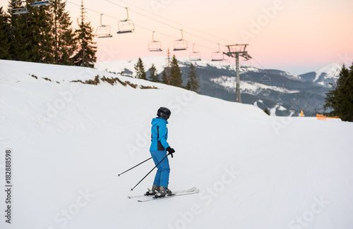 Female skier on the middle of ski slope with ski lifts in background. Woman at ski resort wearing helmet, blue ski suit and goggles. Carpathian Mountains, Bukovel, Ukraine