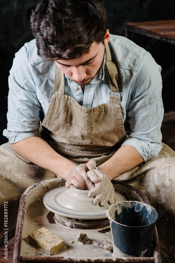 pottery, workshop, ceramics art concept - man working on potter's wheel with raw clay with hands, a male brunette sculpt a utensils near wooden table with tools, master in apron and a shirt pottery, workshop, ceramics art concept - man working on potter's wheel with raw clay with hands, a male brunette sculpt a utensils near wooden table with tools, master in apron and a shirt