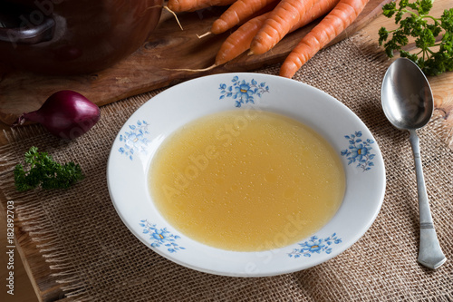 Chicken bone broth in a white plate, with vegetables in the background