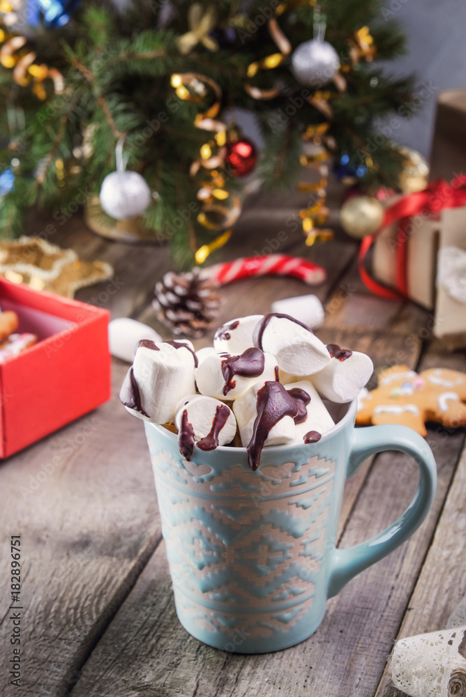 Traditional hot chocolate with marshmallows and chocolate on the old rustic wooden table with christmas tree, decor , gift boxes, cookies and spices. Christmas drink theme. Selective focus.