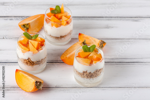 Persimmon creamy trifle in beautiful glasses, fresh ripe fruit slices on white wooden background. Healthy vegetarian food. Delicious dessert. Close up photography. Selective focus.