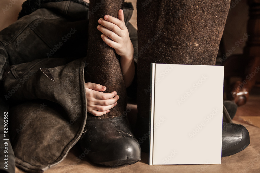 Children's hands protrude from an old coat and embrace dirty felt boots in front of which there is a book