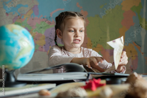 The girl is sitting at the table and angrily unfolds a folded sheet of paper. There is a globe on the table.