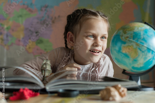 Girl sits at the table and happily looks at the globe. In front of her is a paper boat in an open book