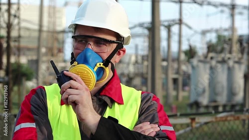 Worker in respirator and with portable radio transmitter on electric power station. Power engineering specialist in gas mask, goggles and hard hat on heat power plant communicates via radio station