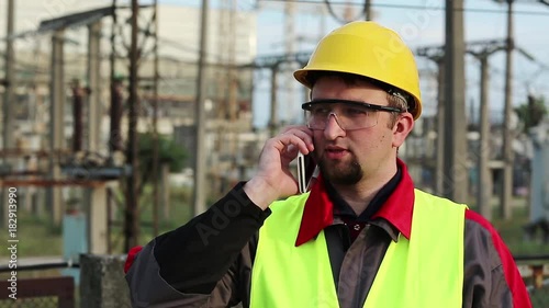 Service engineer at power plant speaks on smartphone. Worker talks on cell phone at heat electric power station near outdoor switchgear. Powerman in yellow hard hat communicates via mobile phone photo