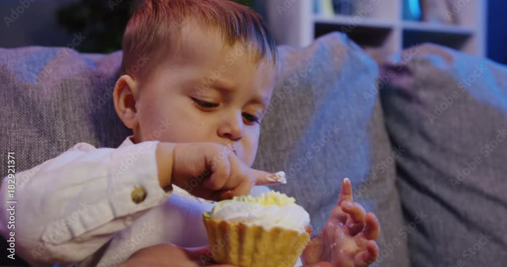 Close up of the funny little boy eating cream from the cake by a finger on the couch in the living room. Portrait. Inside Close up of the funny little boy eating cream from the cake by a finger on the couch in the living room. Portrait. Inside