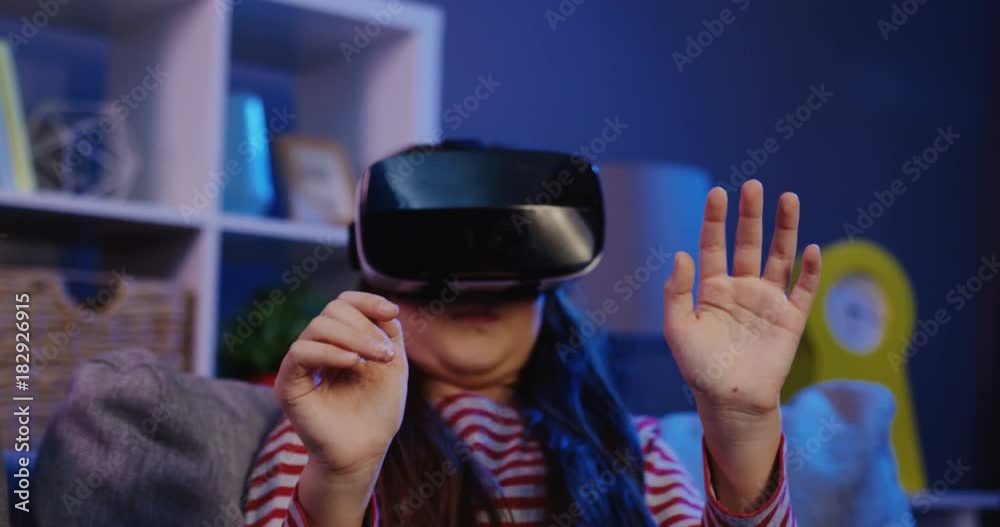 Close up of the schoolgirl with long dark hair having a VR headset while sitting on the couch in the cozy room at night. Girl in VR glasses. Portrait shot. Indoors Close up of the schoolgirl with long dark hair having a VR headset while sitting on the couch in the cozy room at night. Girl in VR glasses. Portrait shot. Indoors