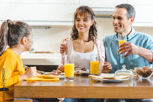 cheerful family sitting at table and holding glasses with juice at kitchen