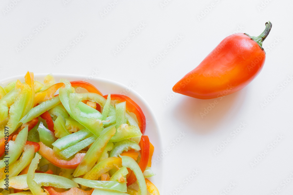 Part of white plate with cuted pieces of colorful peppers and whole uncut orange pepper on white table. Top view. Part of white plate with cuted pieces of colorful peppers and whole uncut orange pepper on white table. Top view.