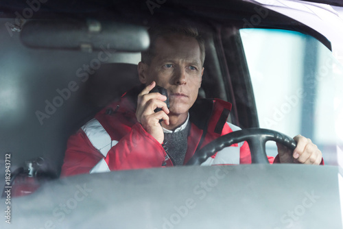 serious paramedic sitting in ambulance on driver seat and talking by portable radio