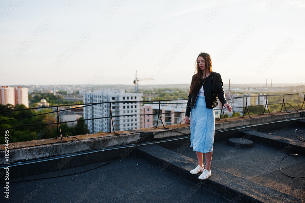 Portrait of a beautiful young woman in black leather jacket and blue dress standing on a rooftop.