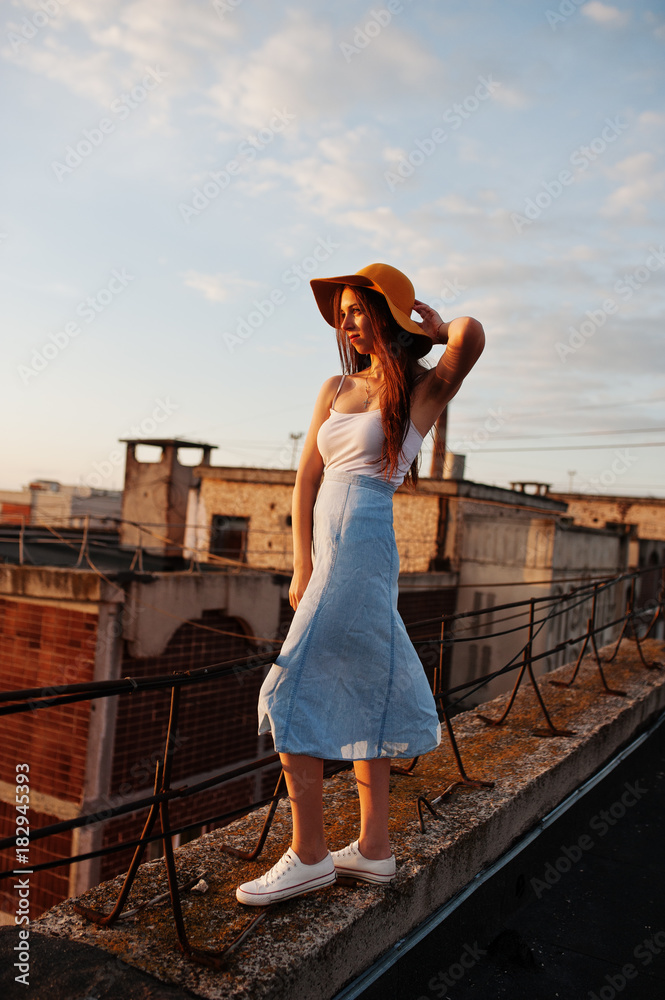 Portrait of a pretty young woman in white t-shirt and blue skirt posing on the rooftop with her orange hat at the sunset.