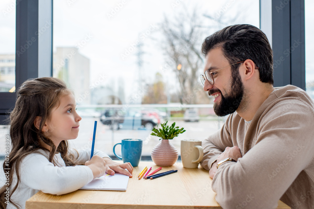 smiling father looking at daughter while she drawing in album