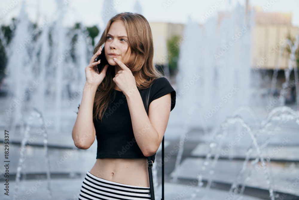Beautiful young woman calling by the phone at the fountain on the street in summer