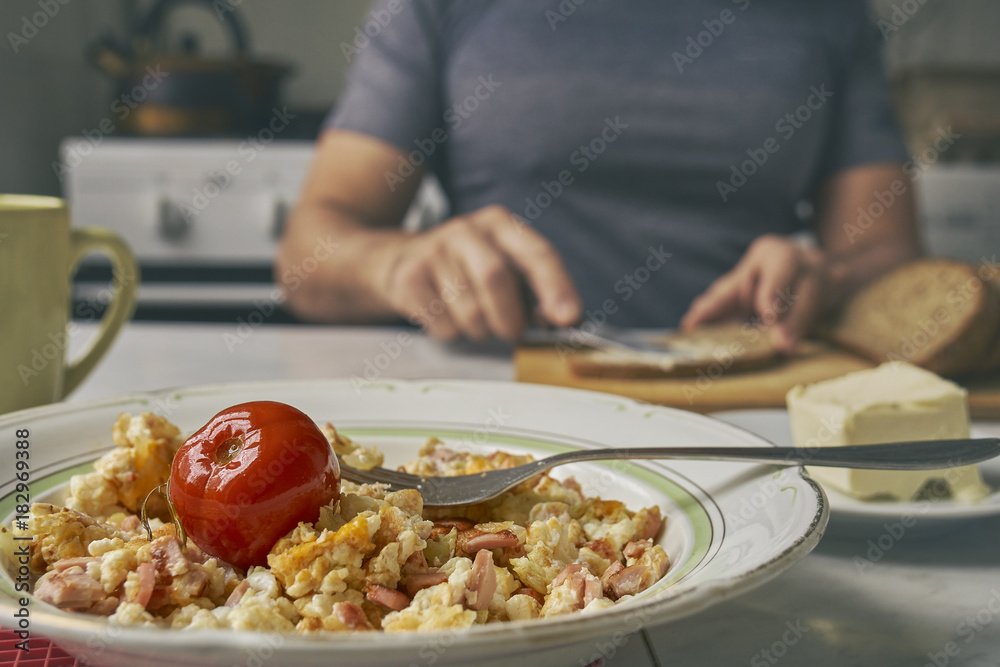 Freshly made omelette with sausages  in a plate on the foreground and a man smearing a butter on bread on blurred background    