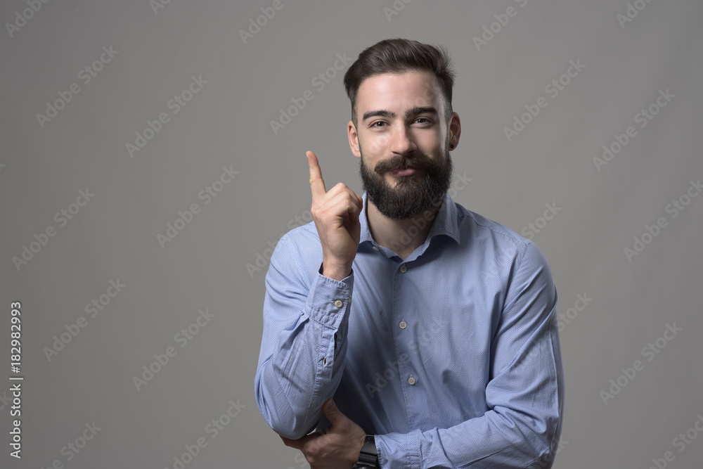 Happy young bearded business man pointing finger upwards smiling and looking at camera with copyspace against gray studio background. Happy young bearded business man pointing finger upwards smiling and looking at camera with copyspace against gray studio background.