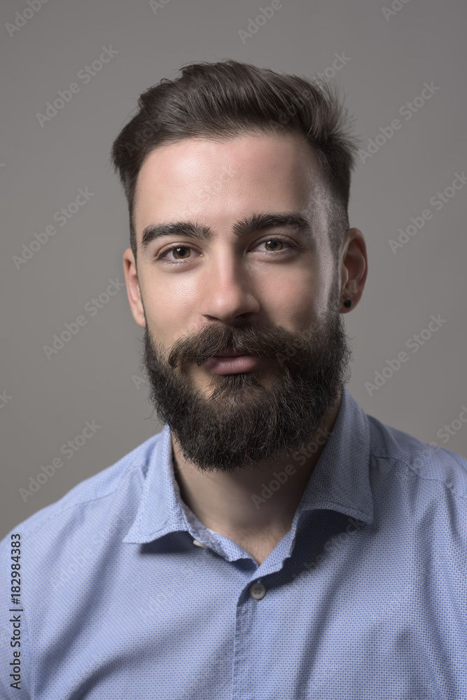 Fototapeta premium Moody bliska portret młodego brodatego mężczyzny, uśmiechając się i patrząc na kamery na szarym tle studio.
