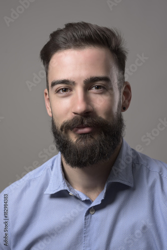 Moody close up portrait of young bearded man smiling and looking at camera against grey studio background. 