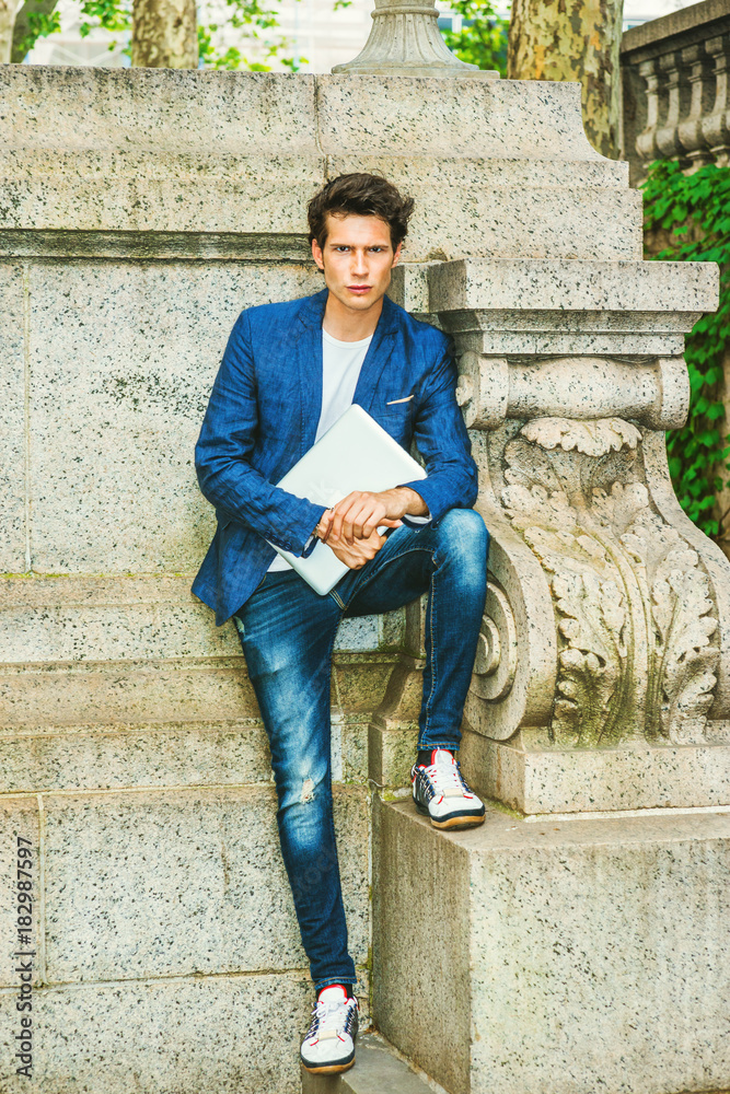 Serious European college student studying in New York. Wearing blue blazer,  jeans, sneakers, holding a laptop computer, a young guy standing at the  corner on campus, waiting for you. . Stock Photo
