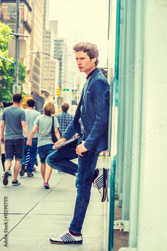 Wearing blazer, jeans, sneakers, holding laptop computer, a young European student standing against glass wall on street in New York, feeling lonely. Young couple holding hands, walking on background