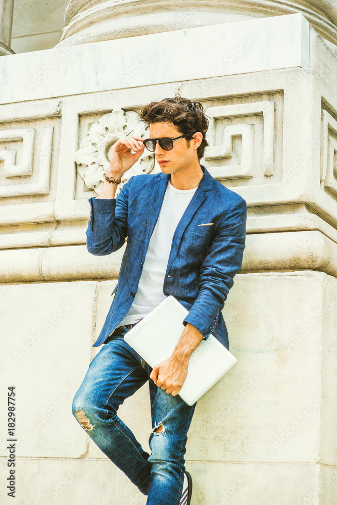 European college student studying in New York. Wearing blue blazer, jeans, sunglasses, holding laptop computer, a young guy standing by column on campus, looking down, thinking. . European college student studying in New York. Wearing blue blazer, jeans, sunglasses, holding laptop computer, a young guy standing by column on campus, looking down, thinking. .