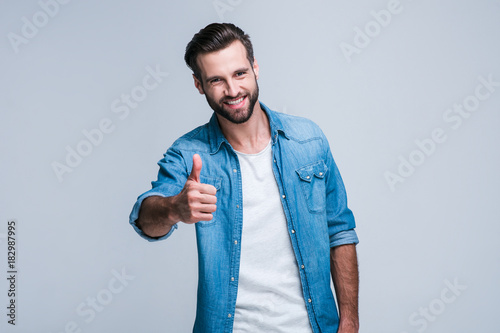 Great work! Handsome young man looking at camera with smile and gesturing thumb up while standing against white background