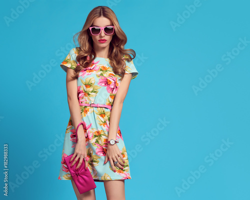 Fashion. Glamour Sexy Blond Model in fashion pose. Young woman in Floral Dress. Trendy, Stylish wavy Hairstyle, fashion Sunglasses, Pink Clutch. Playful Summer Girl