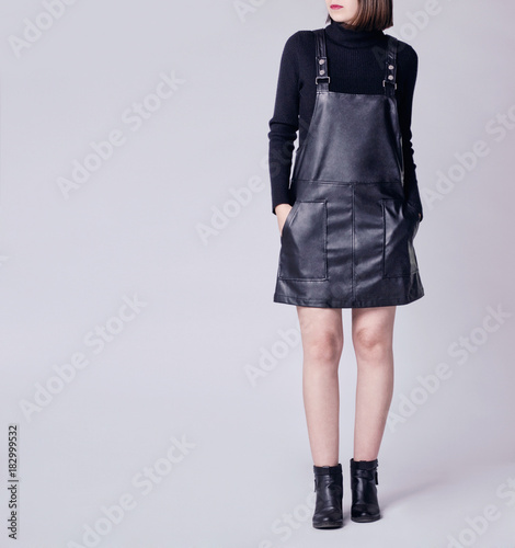 Young woman wearing stylish outfit with black faux leather pinafore dungaree  mini dress, black turtleneck and black ankle boots isolated on light  background. Copy space. Fashion details Stock Photo | Adobe Stock