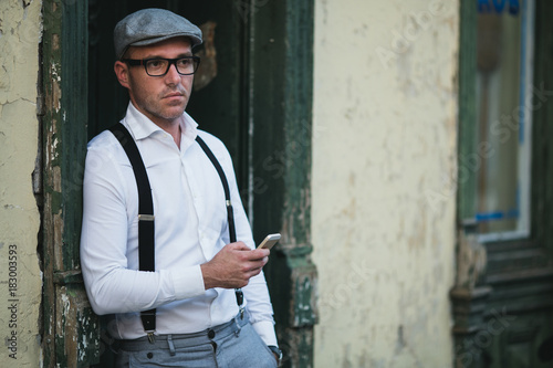 Fashionable retro dressed man with cap, suspenders and eyeglasses standing on city street and using smart phone.