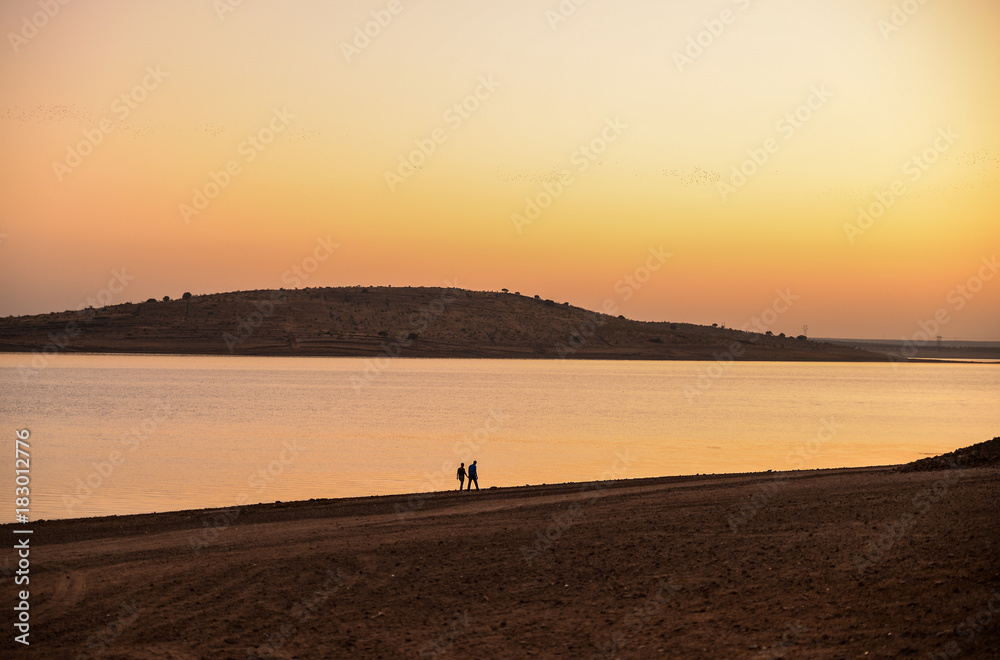 Beautiful view of couple walking together at beach of lake on sunset. Romantic spanish sky, romantic time together. Orange and violet horizon. Beautiful view of couple walking together at beach of lake on sunset. Romantic spanish sky, romantic time together. Orange and violet horizon.